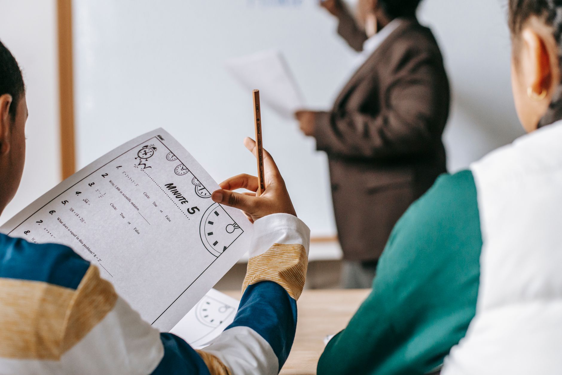 diverse crop pupils sitting at desk against black teacher